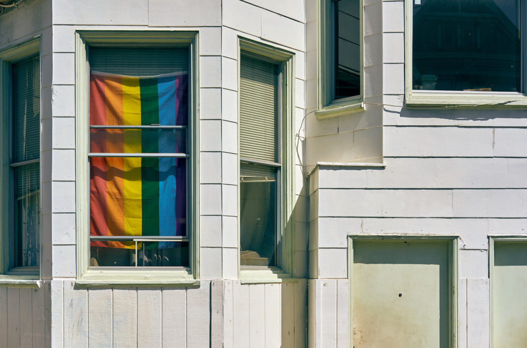 Den officiella regnbågsflaggan skymtar bakom en fönsterruta i ett amerikanskt hem med vit vägg.