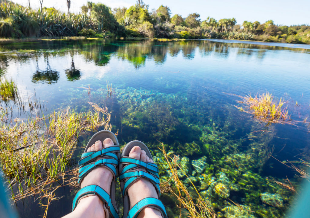 Turkosa sandaler på fötter i nedre delen av bilden. En bit bort ett vattendrag och gröna träd en fin sommardag med blå himmel.