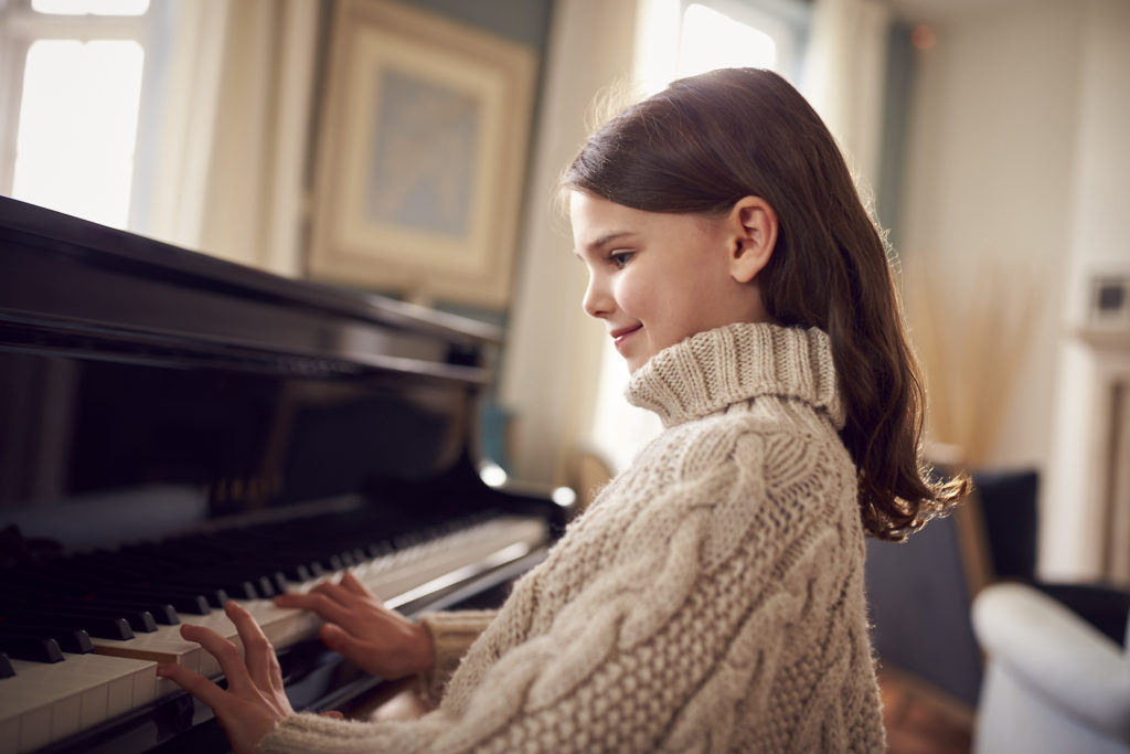 En liten flicka med långt brunt hår och stickad beige tröja intill ett piano. Hon ser glad ut. I bakgrunden syns diffus inredning i ett elegant trevligt hem.