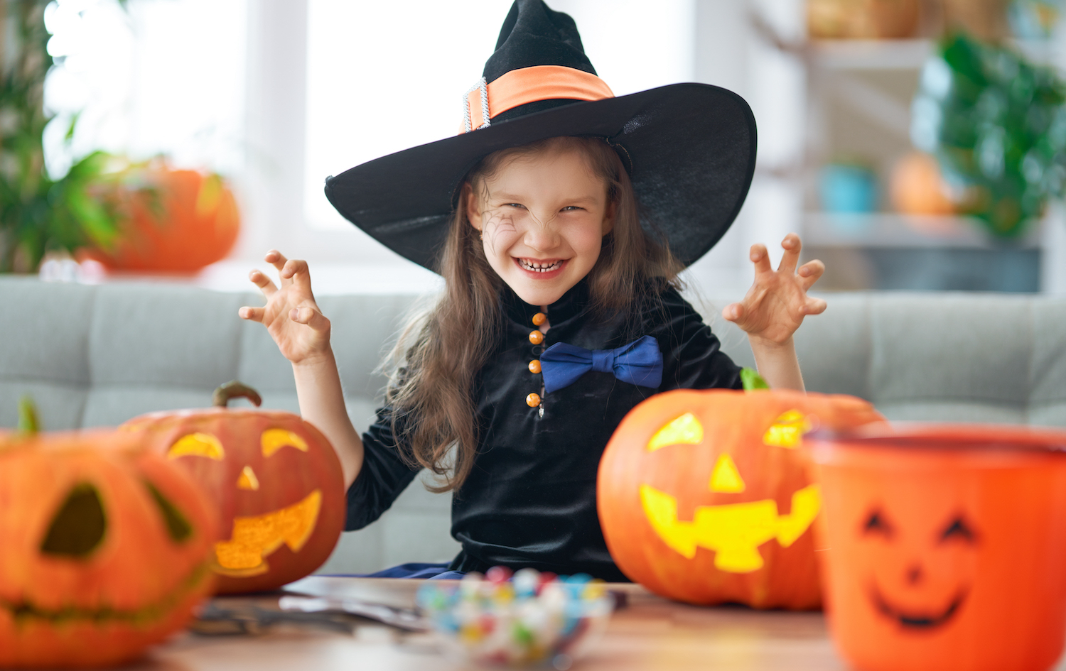 Genrebild; foto på busigt barn som är utklädd till häxa. Hon står och grimaserar i svarta kläder och stor hatt med halloweenpumpor i orange färg på bordet. 