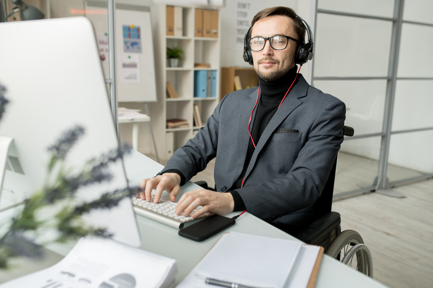 En välklädd korthårig man i mörk kostym och glasögon sitter vid skrivbord och arbetar på dator. Han är uppkopplad till telefonen med headset och ser produktiv ut. I bakgrunden syns whiteboardtavla och bokhyllor.