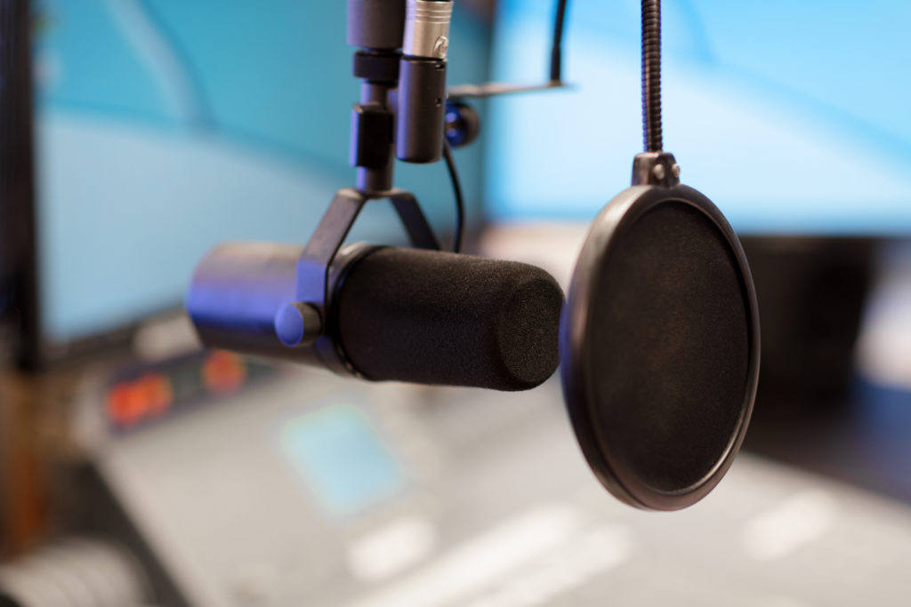 En svart mikrofon i närbild fotad i radiostudio med skärmar och mixerbord suddigt i bakgrunden.