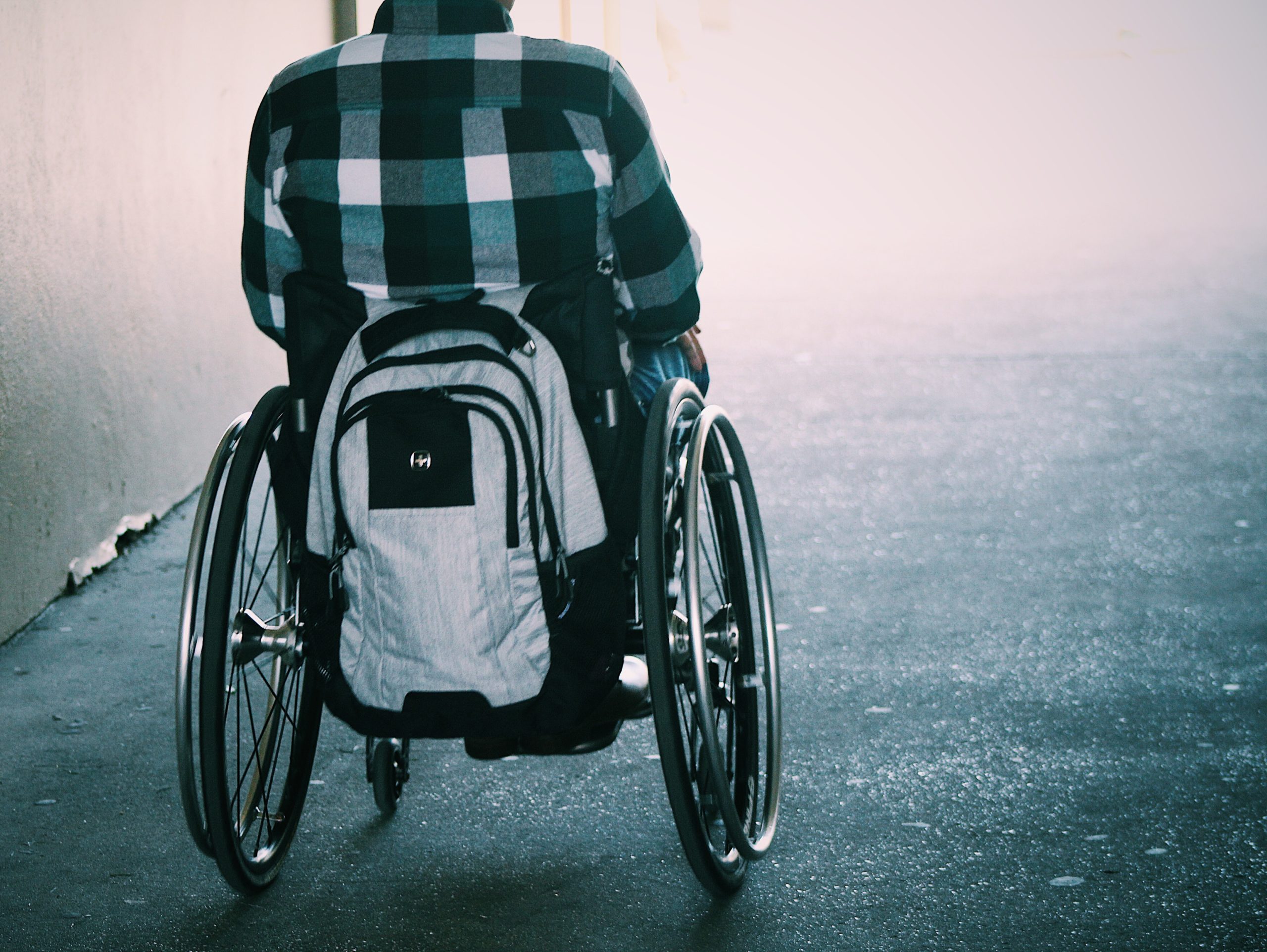 Ryggtavla av en person i rullstol. Bak på rullstolen hänger en vit ryggsäck. Personen har en vitblårutig skjorta och bakgrunden är suddig med ljus. Hela bilden har en blåaktig ton.