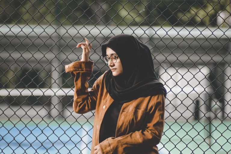 Kvinna med hijab står vid stängsel. Hennes hijab har mörk färg, hon bär glasögon och en ljusbrun jacka. Hon funderar på något och ser något bekymrad ut.