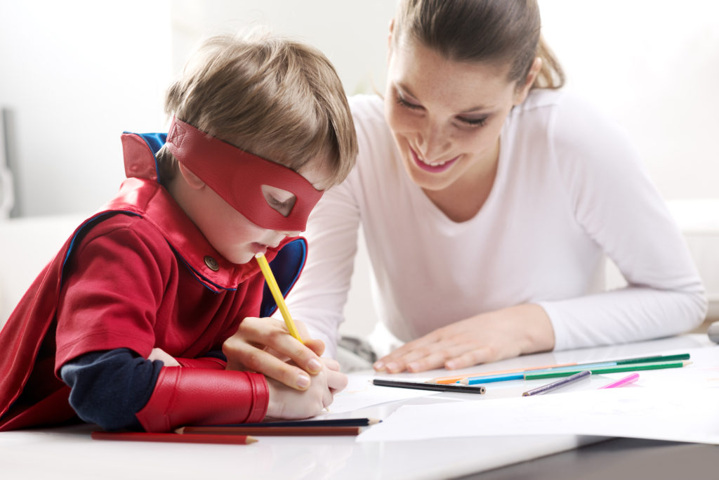 Ett barn sitter med vuxen vid bord och lär sig rita med hjälp av pennor och papper. Han har på sig en rör superhjältedräkt och mask lekfull mask som täcker runt ögonen.