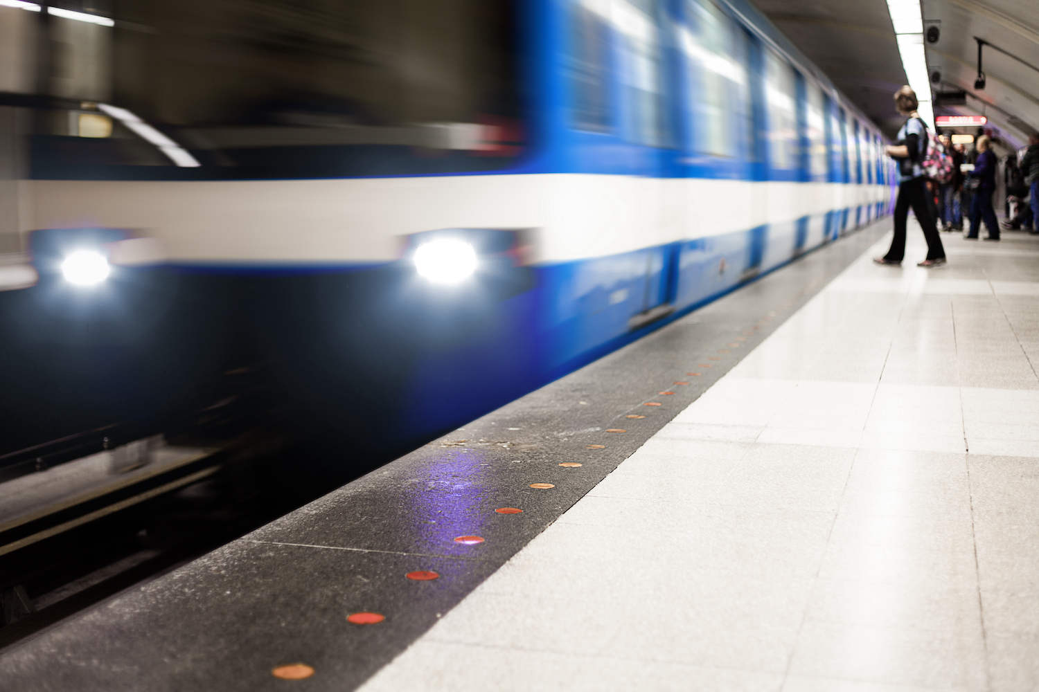 Ett blått tåg i rörelse intill perrong där människor väntar på att tåget ska stanna.