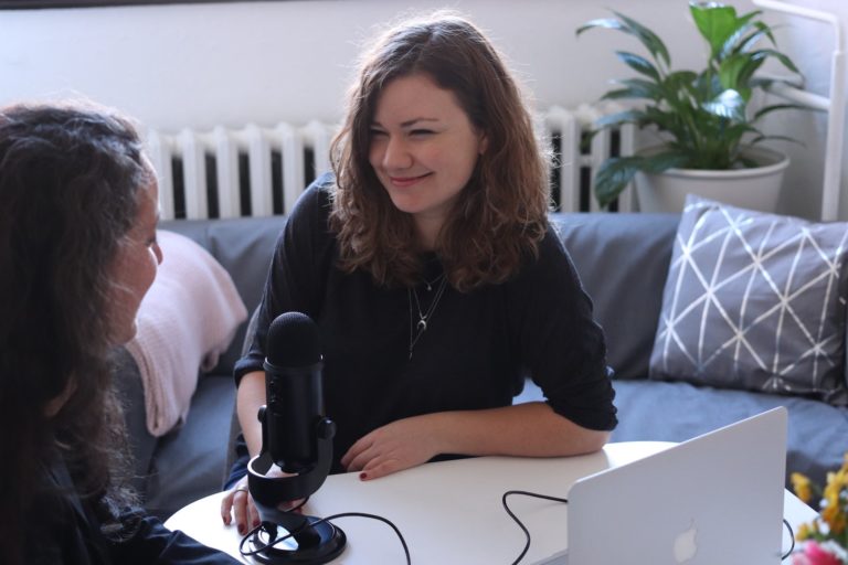 Två tjejer sitter i intervjusituation med en bordsmikrofon och dator emellan dem. De har brunt långt hår och mörka kläder. I bakgrunden syns en blå soffa och vit vägg.