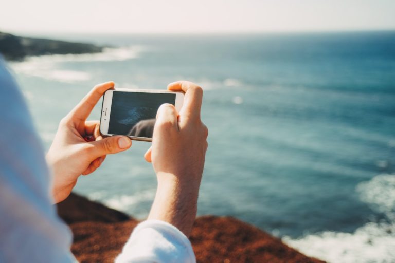 Två hände håller om en mobiltelefon och tar foto på vågorna från stranden på en resa