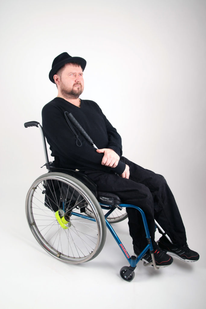 Petri Mykkenän sitter i rullstol i fotostudio med vit bakgrund. Han har på sig mörka kläder och en svart hatt. Han har kort skägg och en käpp i handen.