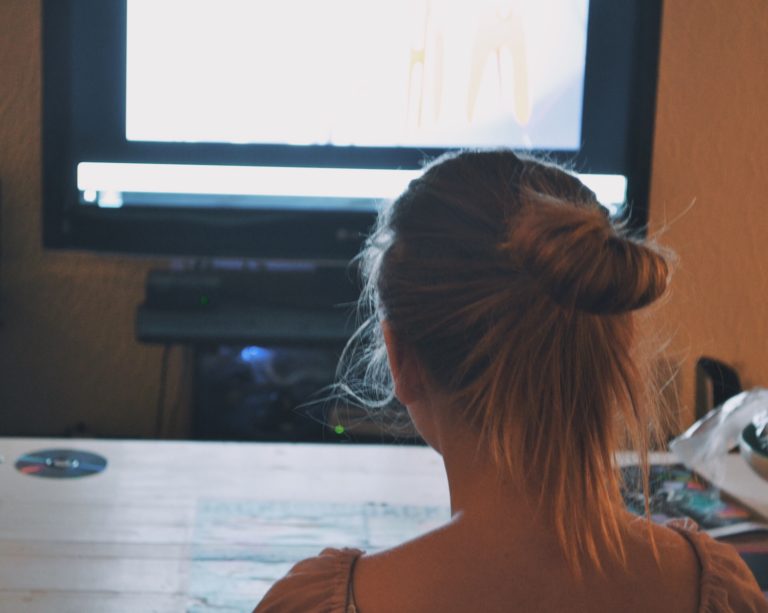 En tjej i ljust långt hår fotad bakifrån, tittar mot en tv-bild
