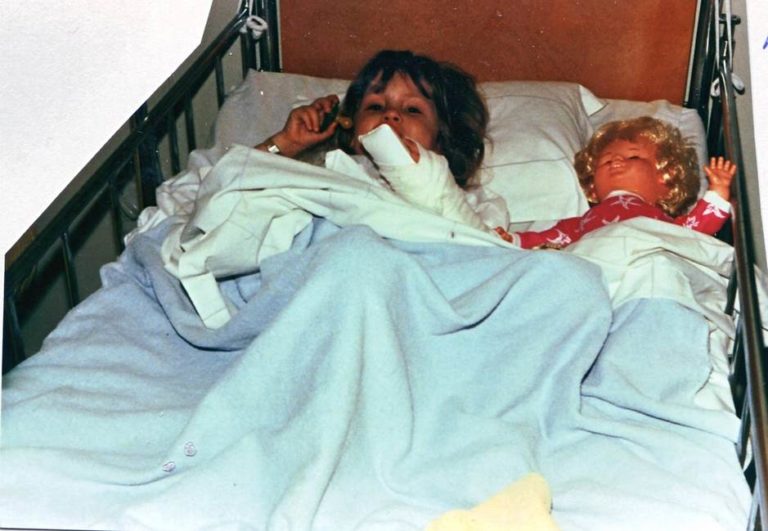 Jag som barn på sjukhuset. Jag ligger i en barnsäng intill en docka. Vi ligger båda under ett ljusblått täcke. Jag har mörkblont hår och dockan blont hår.