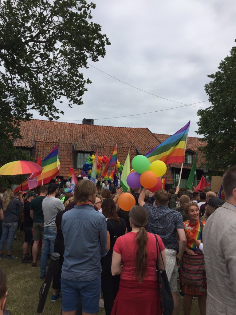 Foto från Mångfaldsparaden i Almedalen. En stor grupp människor viftar med regnbågsfärgade balonger och flaggor på ett fält intill ett hus. 