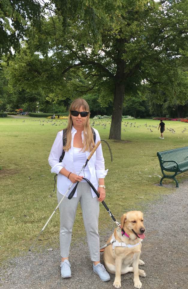 Foto på mig och Chanti promenerandes på Djurgården. Jag har på mig vit skjorta, glasögon och ljusgrå byxor och skor. Chanti är en ljus labrador som bär ett svart koppel. I bakgrunden syns en grön park en somrig dag.