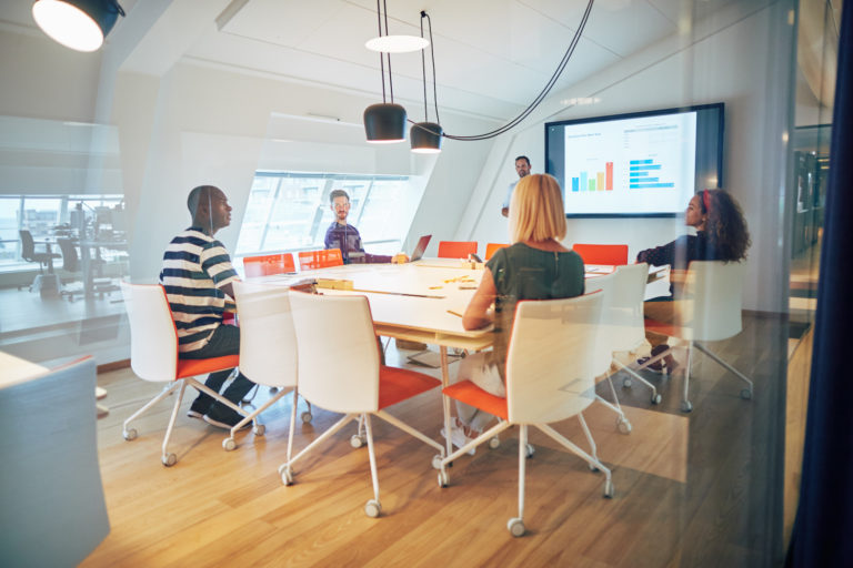 En arbetsgrupp med olika kön och olika etnisk bakgrund samtalar på kontor. samlat kring ett mötesbord. En person står och visar grafer på en storbildsskärm.