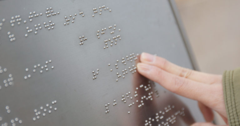 Två fingrar känner sig fram över en silverfärgad metallplatta som innehåller punktskrift