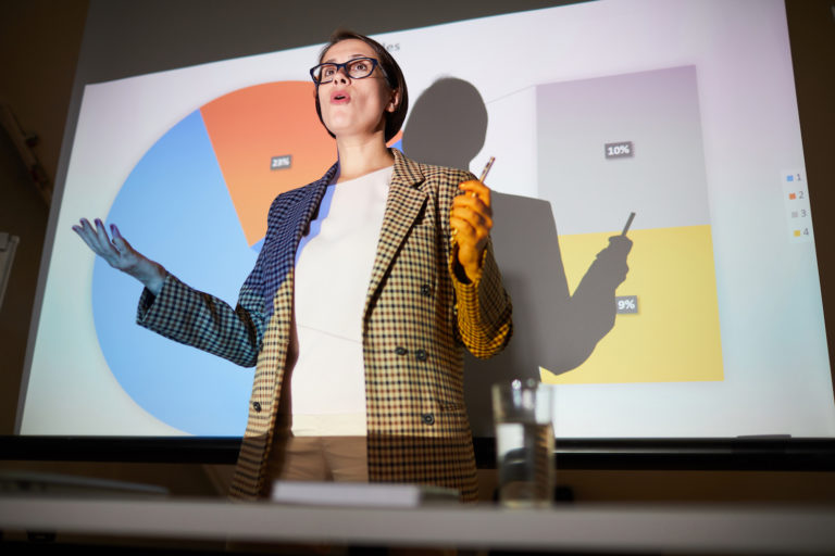En kvinna med brunt kort hår föreläser nära en upplyst projektorduk. På duken syns olika procenttal i någon form av statistisk illustration.