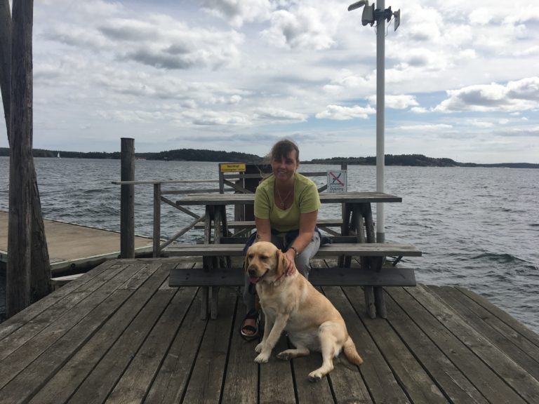 Jag med min ledarhund Chanti. Vi sitter en sommardag på en bänk intill vattnet på ett trädäck.