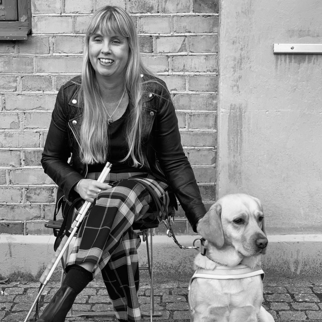 Me sitting dressed in black leather jacket, next to me my cream colorued guide dog wearing a white harness.