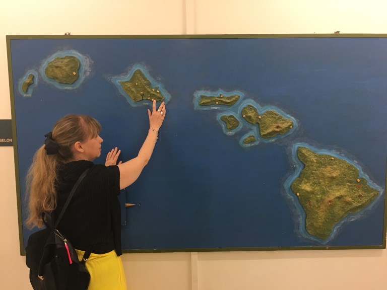 Jag håller handen på en taktil kartan över Hawaii. Kartan har blått vatten och gröna öar. Jag bär svart tröja och gula byxor.