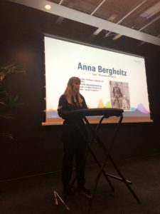 Jag föreläser vid ett ståbord på en scen, iklädd svart byxdress. På en skärm i bakgrunden syns en bild på mig där jag sitter med min ledarhund och rubriken "Anna Bergholtz".