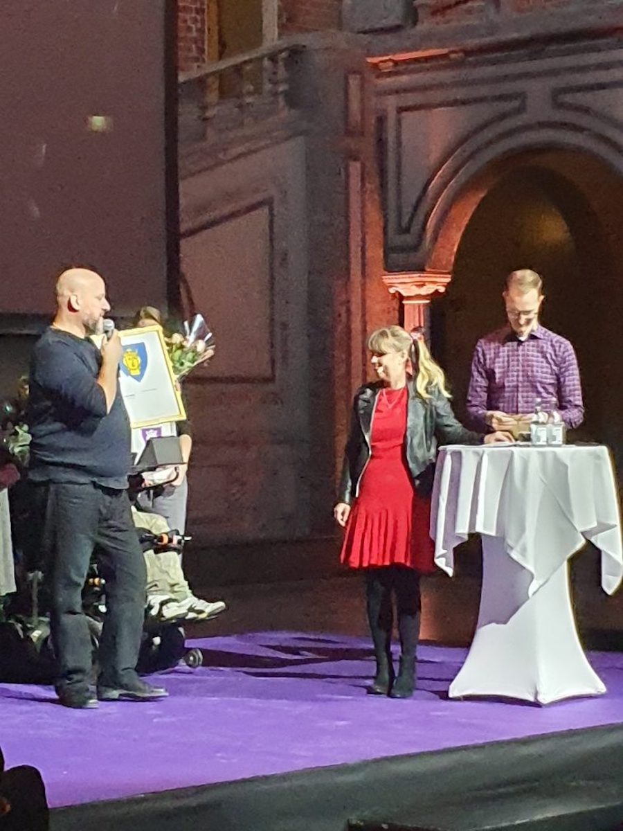 Jag på scen vid vitt ståbord, i röd klänning under svart skinnjacka. PÅ scen även Jan Jönsson och Håkan Leydner.